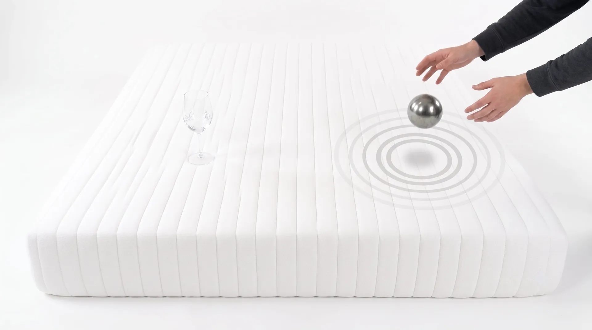 Hands dropping a weighted ball on a white quilted Twin XL mattress while a glass of wine stands on the other side